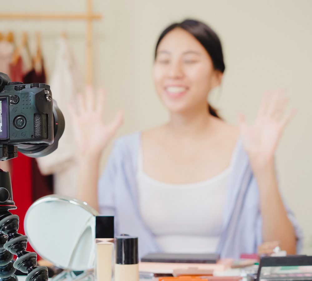 Beauty blogger present beauty cosmetics sitting in front camera for recording video. Happy beautiful young Asian woman use cosmetics review make up tutorial broadcast live video to social network.